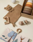 Flat lay of modern baby toys including a lovey, silicone stacking tower, wooden rattle, teether, and woodland sensory book in warm neutral tones.