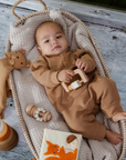 Baby lying in a cozy basket surrounded by neutral baby toys including a wooden rattle, lovey, and silicone stacking tower.
